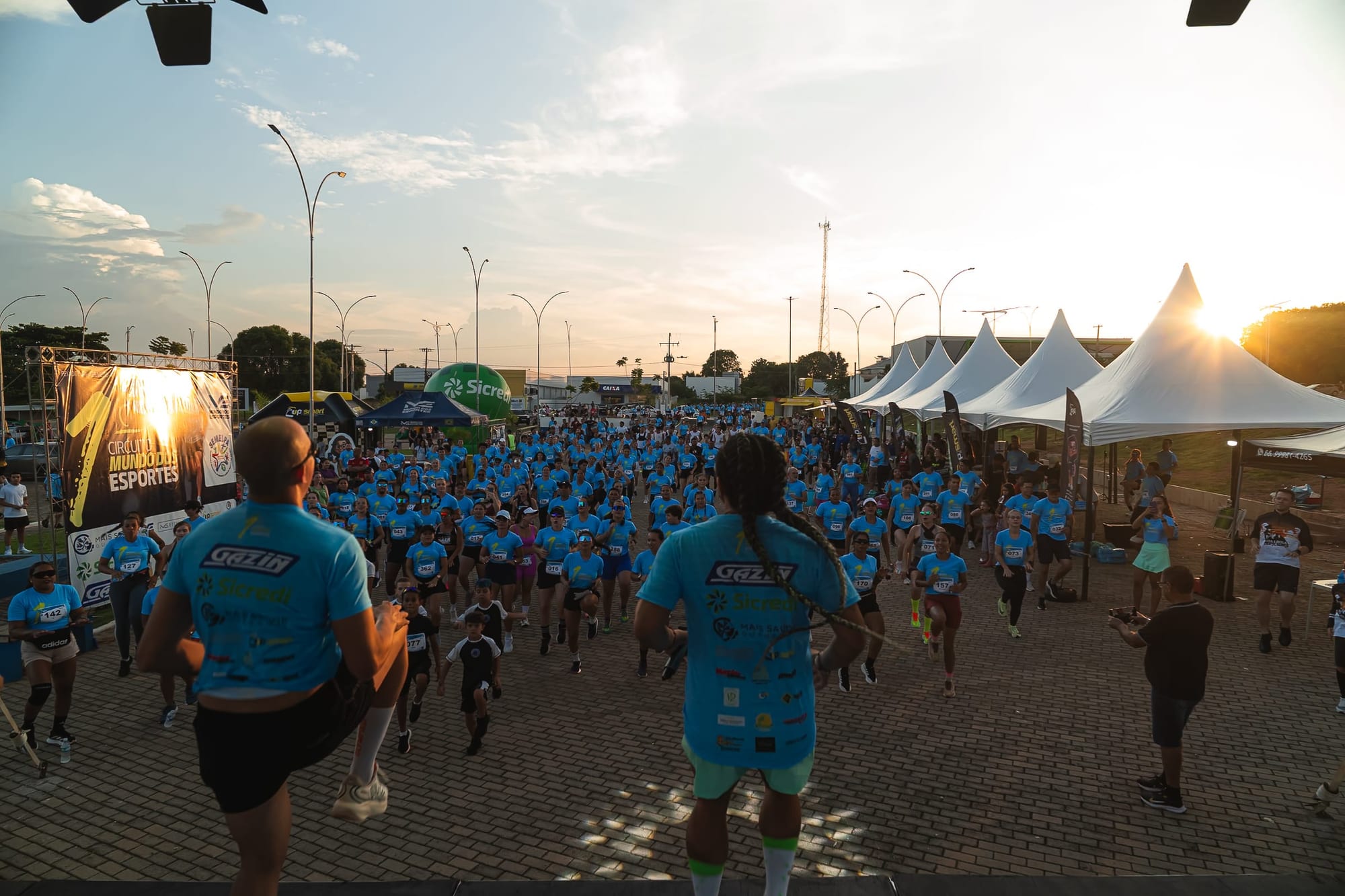 Centenas de atletas de camisa azul no aquecimento da 1ª edição, ao pôr do sol, com o palco e as tendas ao fundo